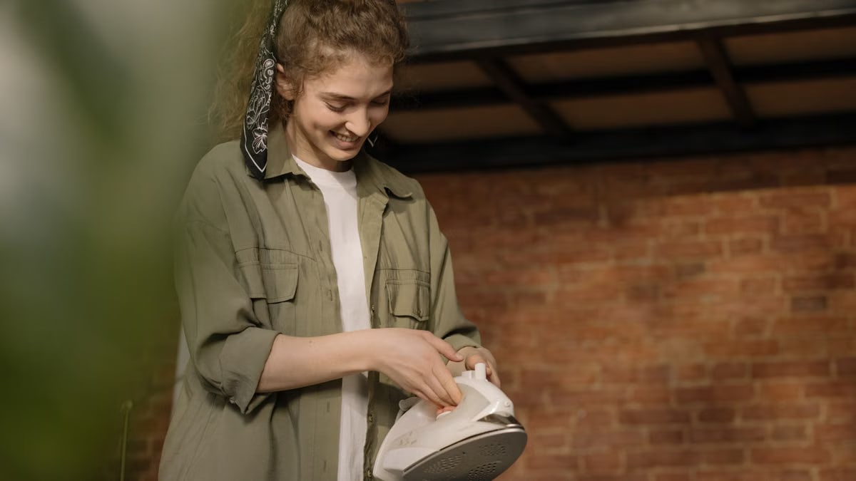 A young woman with curly hair ironing clothes in a cozy, modern home interior with a brick wall.